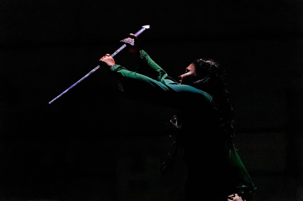 Female dancer in dark green shirt holding up a sword in dramatic spotlight