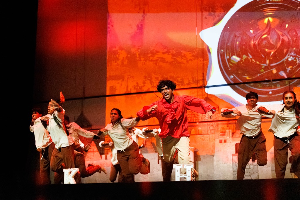 Lead dancer in red shirt running on stage with dancers posing behind
