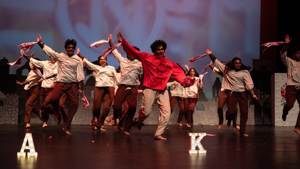 Lead dancer in red shirt running/jumping mid-air with other dancers in background